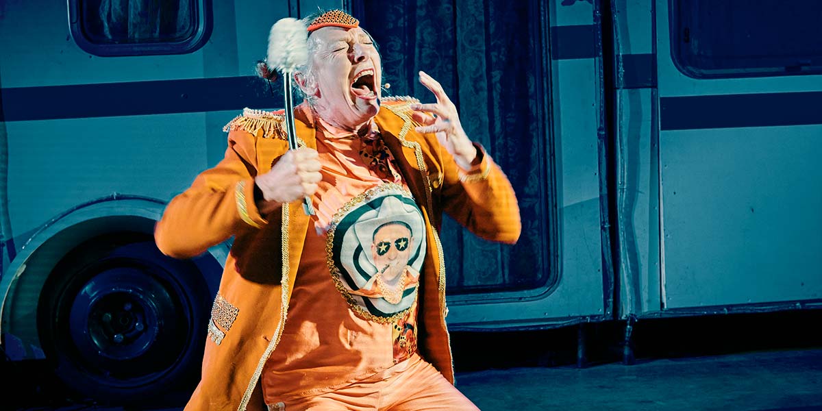 Image description: A performer dressed in orange kneels on the floor, excitedly wailing. She clutches a toilet brush in one hand. On her t-shirt is the cartoon image of a man in a Stetson hat.