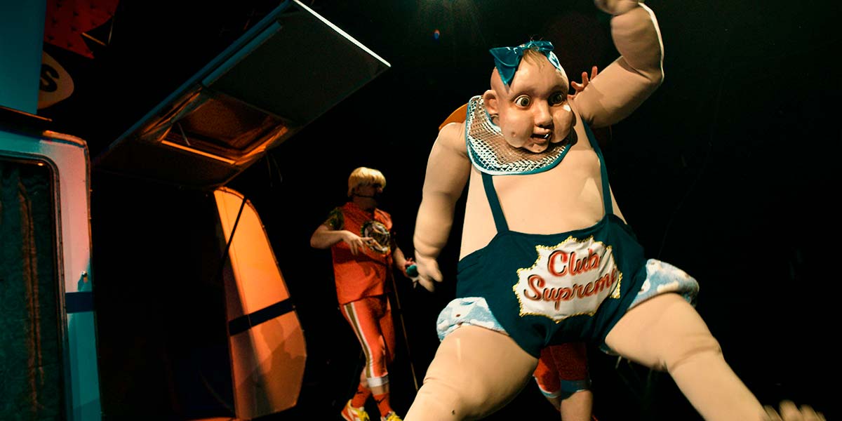 Beneath a bright theatre light, a giant puppet baby holds one arm in the air and stares blankly through its wide eyes. It wears a blue nappy with the Club Supreme logo on it.