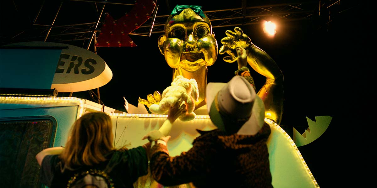 A giant golden baby erupts out of the top of a caravan. It holds one arm in the air whilst two people stand before it, waving a cuddly toy.
