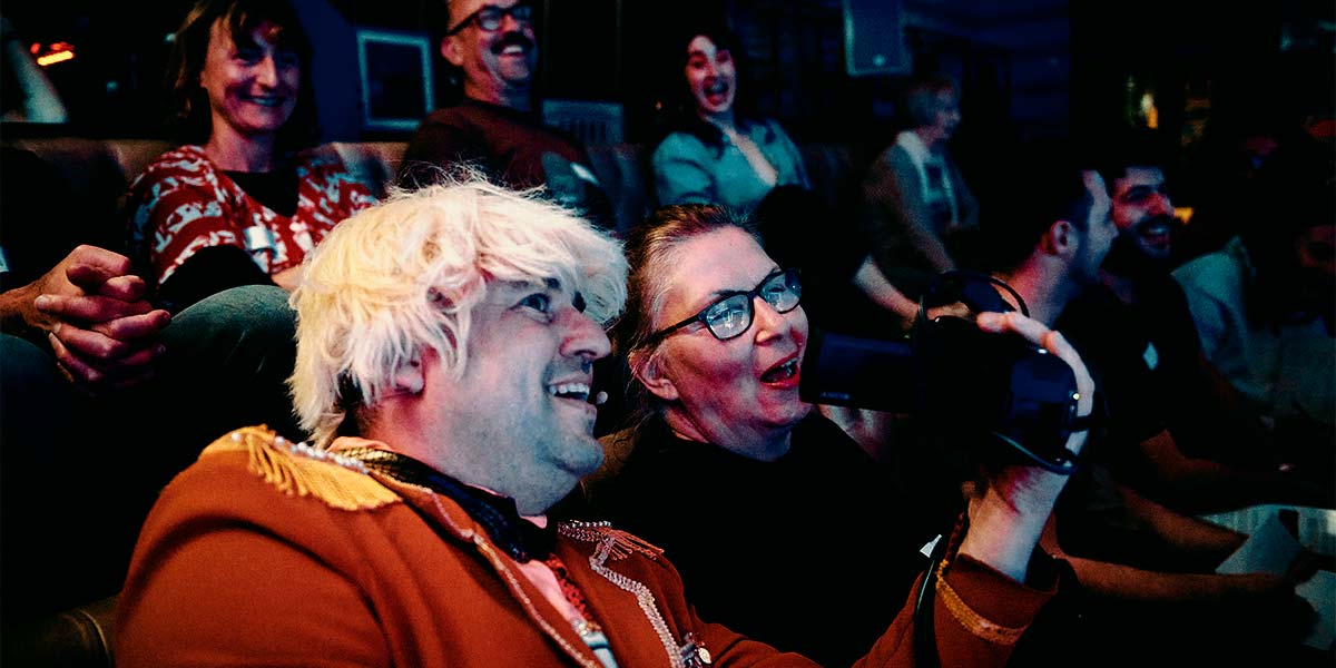 : A grinning man with a bright blond wig, sits amongst a laughing audience. He holds a video camera in his hand and shows the screen to a smiling audience member next to him.