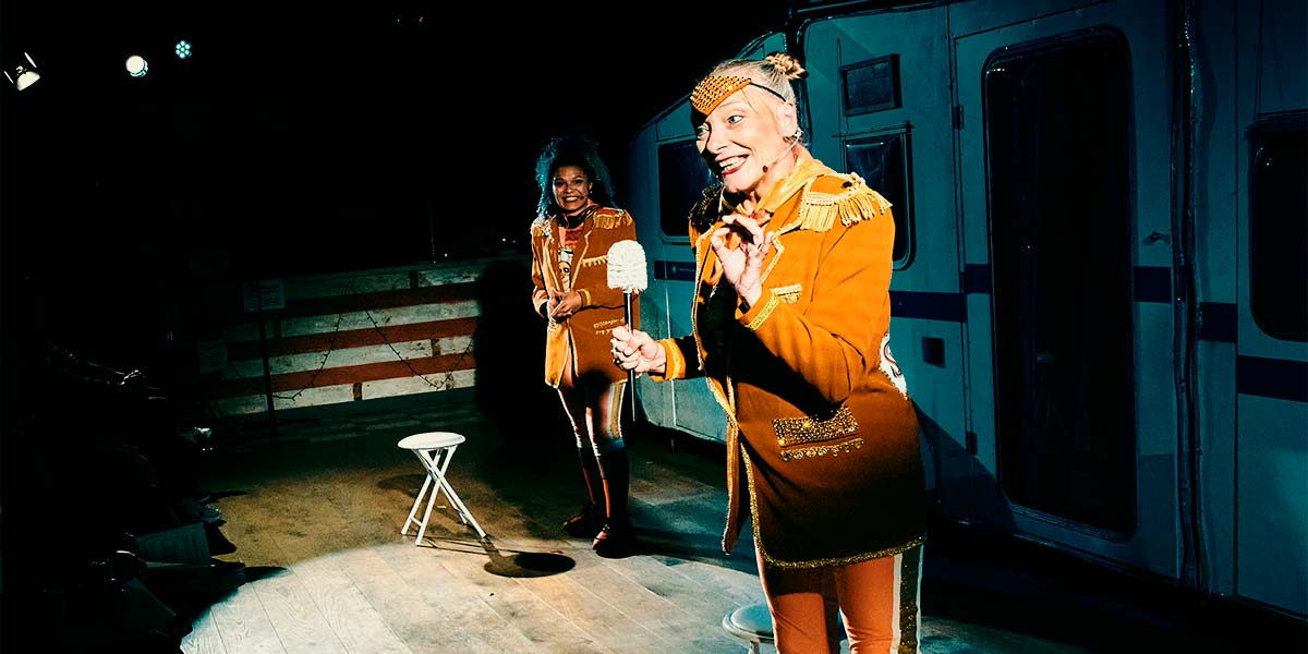 A woman dressed in the Club Supreme uniform of orange leggings and blazer, holds a toilet brush in one hand and grins toothily at the audience sat in front of her. Behind her, another Club Supreme member watches, smiling.