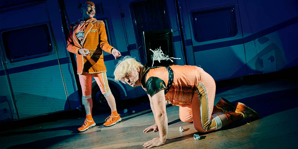  A man with bright blond hair and dressed in orange, kneels on all fours. Balanced on his back is a paper shredder, with paper spilling out of it. To the side of him a woman, also dressed in orange, holds the lead of the shredder and laughs.