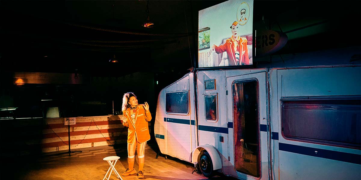 A woman wearing orange, gazes up at a huge screen fixed over the top of a caravan. It shows the image of another woman dressed in the same attire. She sits pertly on a couch and holds up the back of her hand, as if to be kissed.