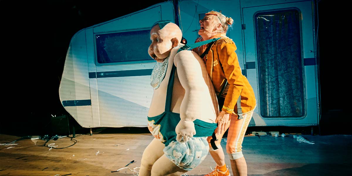 A huge, adult-sized puppet baby is strapped to the front of a Club Supreme performer, whose mouth is open in a wail.