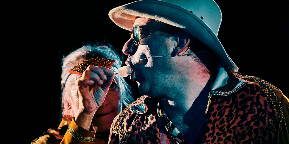 A man wearing a Stetson hat and leopard-print blazer, sucks on a cocktail stick of cheese and pineapple proffered by a Club Supreme member.