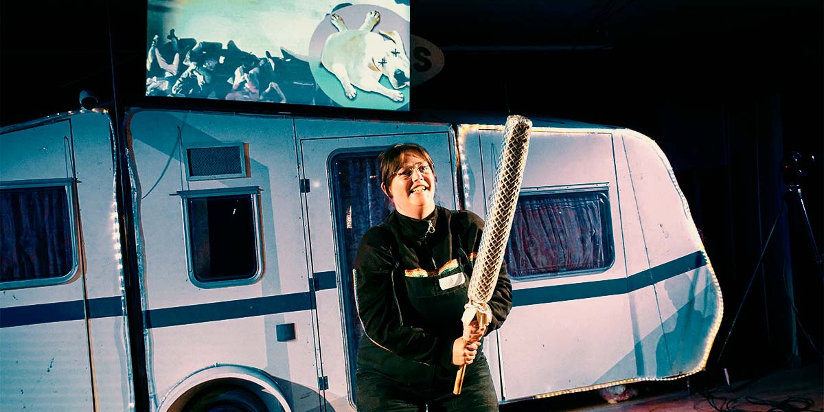 An audience member brandishes a long, foam-covered stick. Behind them is a white caravan, over which a huge screen shows the image of a dead cartoon dog with crosses for eyes.