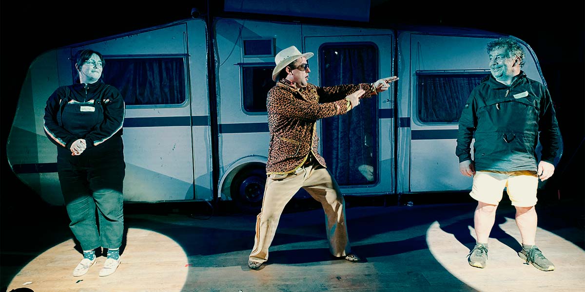 Two audience members are lit by spotlights. A man in a leopard-print jacket and Stetson hat stands between them and gestures to one with double-pointed fingers. The audience member stares back, smiling.