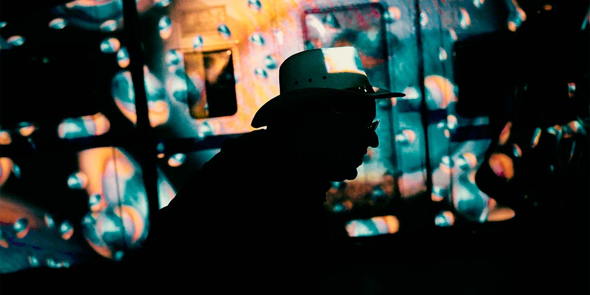A man standing side-on and wearing a Stetson hat is silhouetted against a swirling, spotted background of eyes that are blues, pinks and orange.