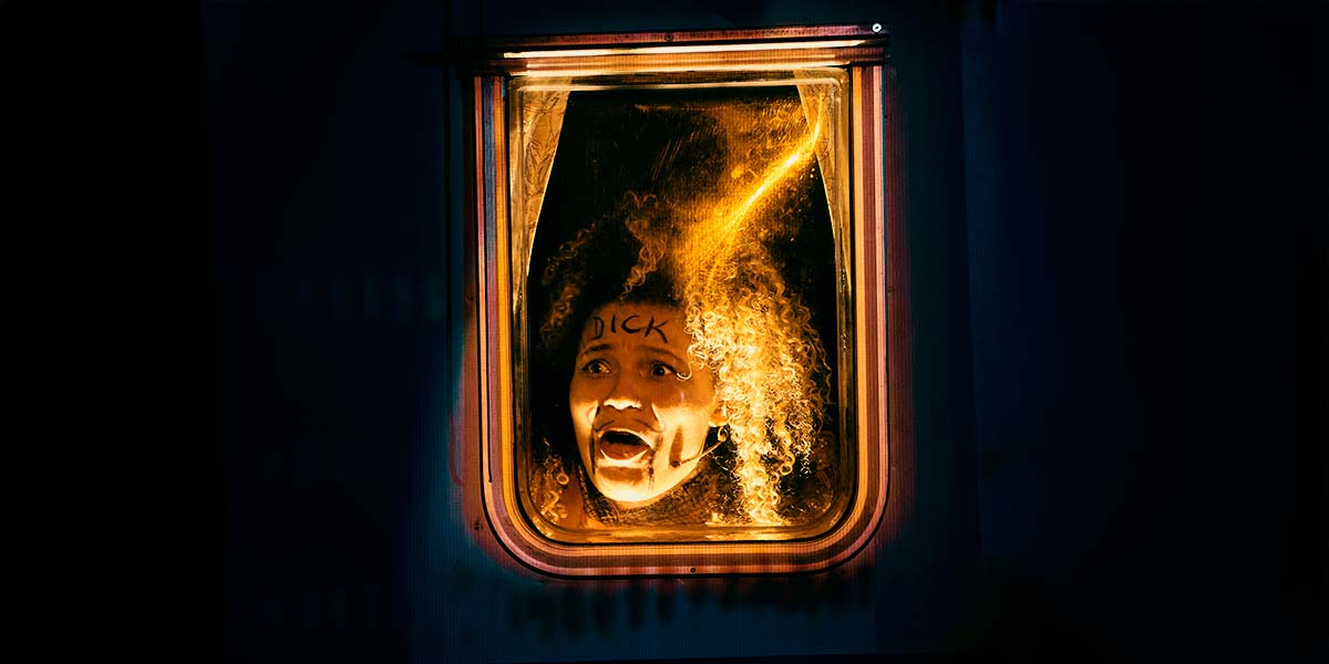 A woman’s face is illuminated behind a neon-edged window. Her eyes are wide; mouth open. She has a fake moustache and beard drawn on her face, and on her forehead the word ‘dick’ is written in capital letters.