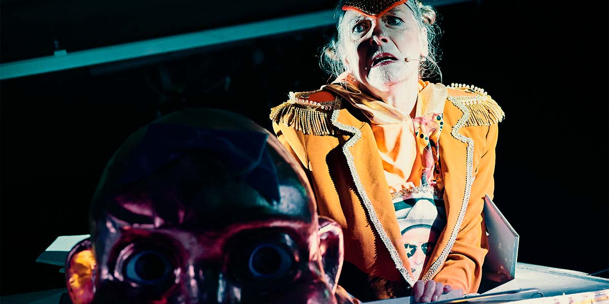 A giant baby’s head glows a fiendish purple. Behind it, a woman emerges through a hatch. She wears an orange blazer with a golden triangle over her brow. She stares out over the top of the baby with wide, fearful eyes and a downturned mouth.