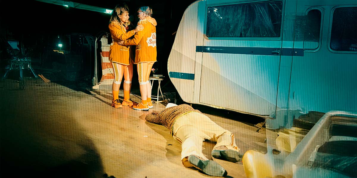 Two women in Club Supreme orange blazers and leggings, grip each other tensely. Sprawled on the floor at their feet, a body lies face down.