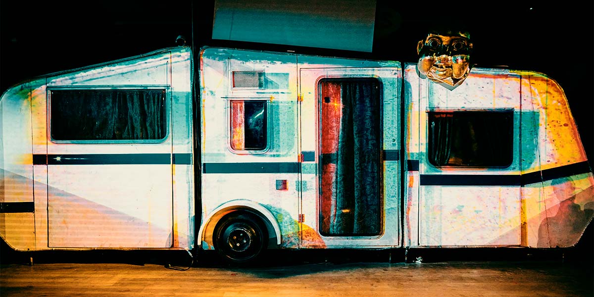 A white caravan sits on a wooden floor, its surface patterned with cool hues of blue and purple light. Dark curtains are drawn behind the windows and door. Above the caravan is a huge blank screen, and over the right-hand window hovers the head of a large, golden baby.