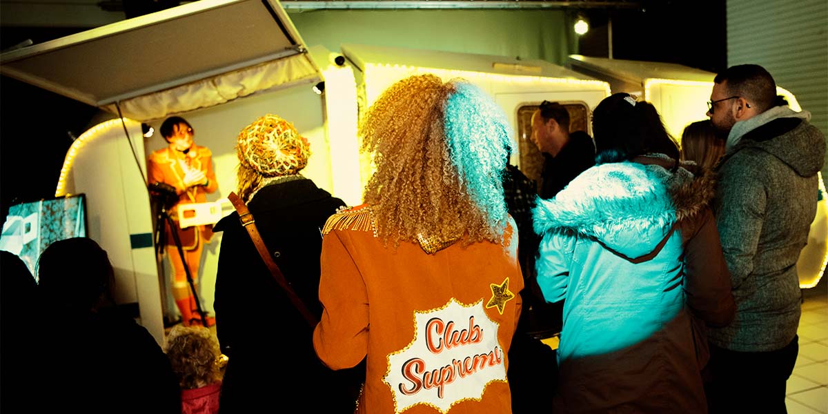 A group of people watch a Club Supreme performer standing inside a caravan. The back of another performer is in focus, with the words Club Supreme emblazoned on her orange blazer