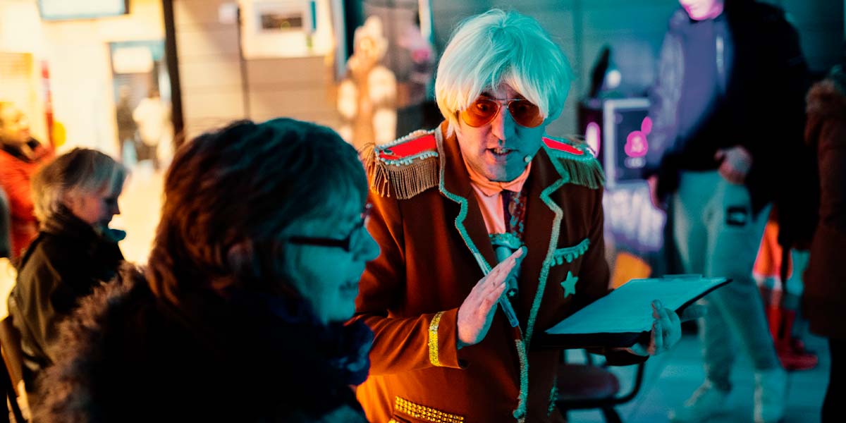 A bystander laughs as a man in the Club Supreme orange blazer and sunglasses approaches her. He has platinum blond hair and holds a clipboard. Behind them, the room is filled with people milling around and smiling.