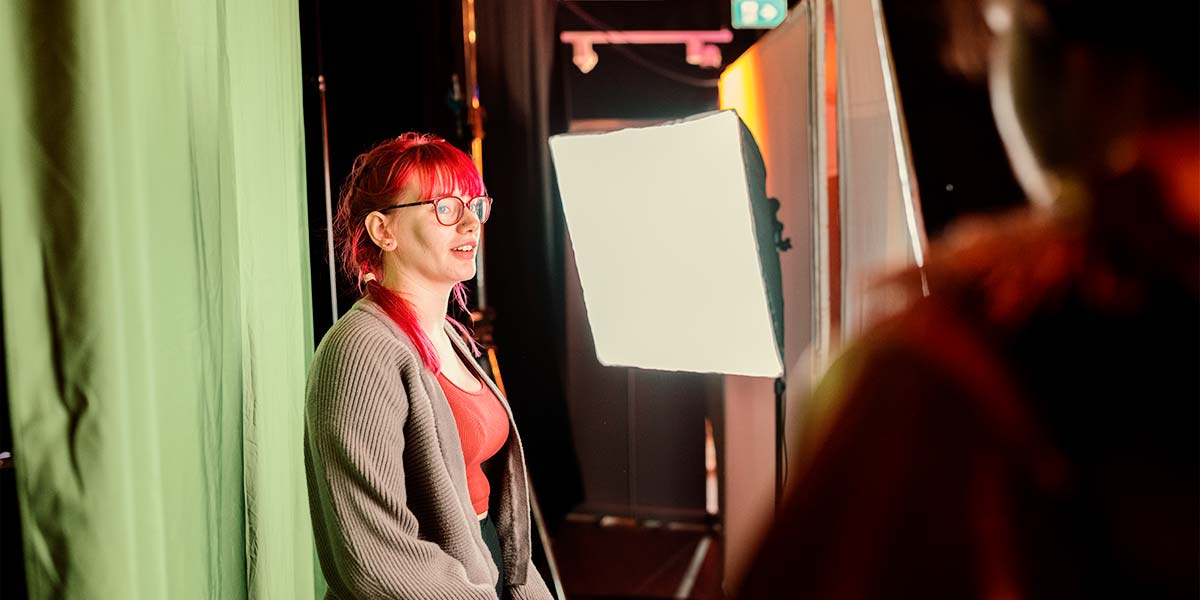 A person with bright pink hair and glasses stands in front of a vivid green curtain. A bright LED screen lights them from the side as they gaze towards an obscured camera.