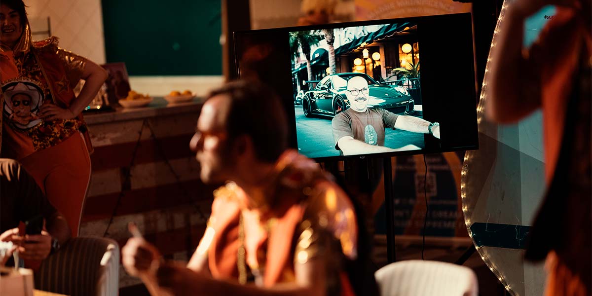 A tv screen shows a grinning person superimposed over the image of a fast car parked outside a hotel. Around the tv screen, the room is crowded with tables and chairs and people talking to each other.