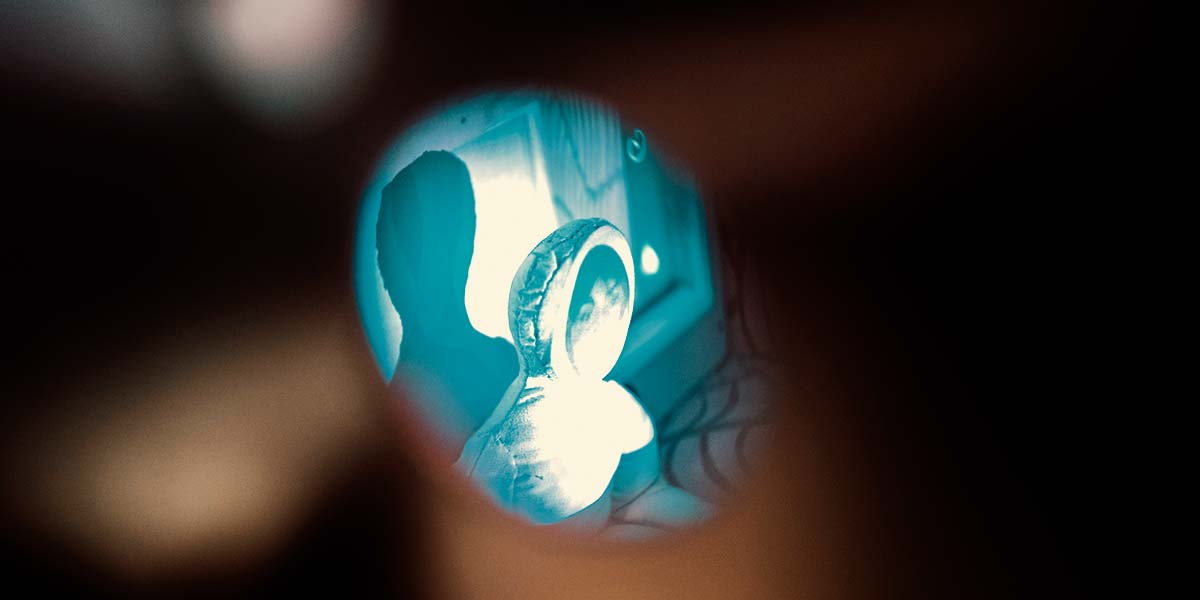 A peephole of light reveals a white rag doll with the face of a person projected onto it.