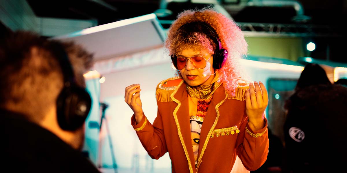 A person with headphones on watches a woman who wears a headset. She is dressed in an orange blazer and holds her hands upwards in a gesture of meditation. Her eyes are closed behind her orange sunglasses.