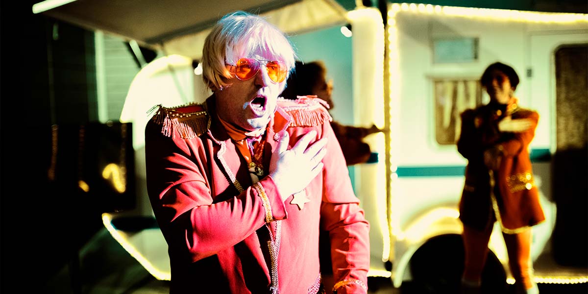 A man stares open mouthed into the distance; his right hand placed over his heart. His orange Club Supreme uniform appears red under the light. Behind him, a green-lit caravan is framed with bright LEDs.