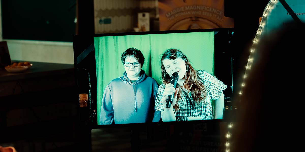 In a room filled with LED lights, Club Supreme posters and paraphernalia, two people appear on a tv monitor in front of a bright green curtain. One smiles tightly, whilst the other sings into a microphone, eyes closed1