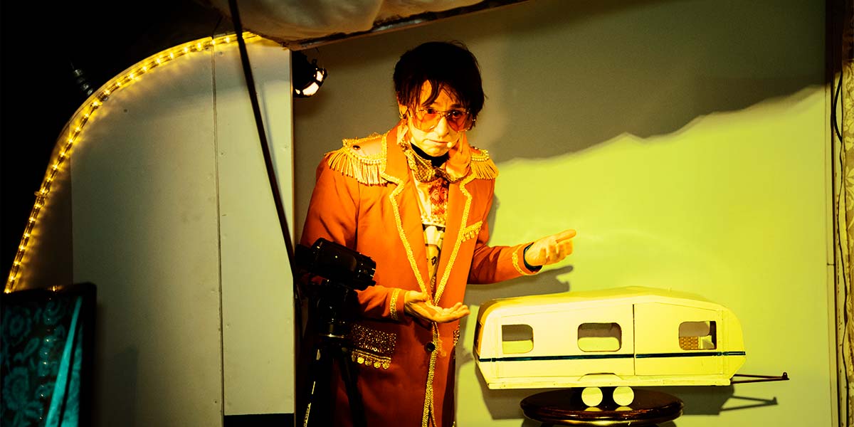 In green light, a woman wearing a Club Supreme blazer trimmed with gold, presents a model of a caravan in front of a small video camera. Her face is solemn as she gazes away from the lens.1
