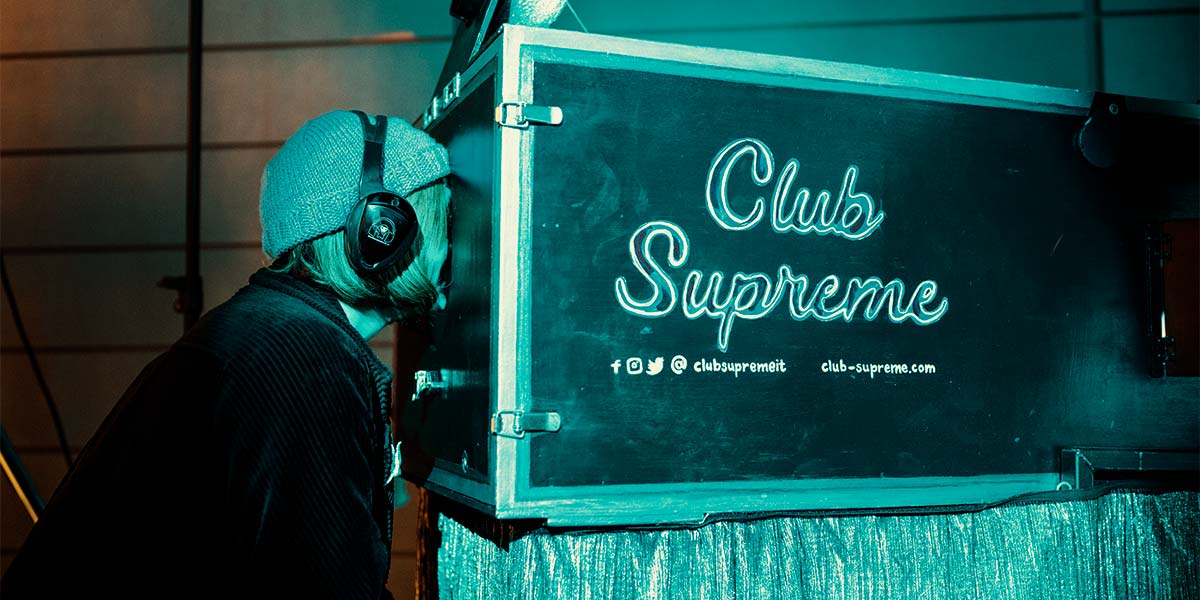 A large, black, rectangular box emblazoned with the words Club Supreme sits on a metallic cloth. A person wearing large headphones looks through a hole in its side