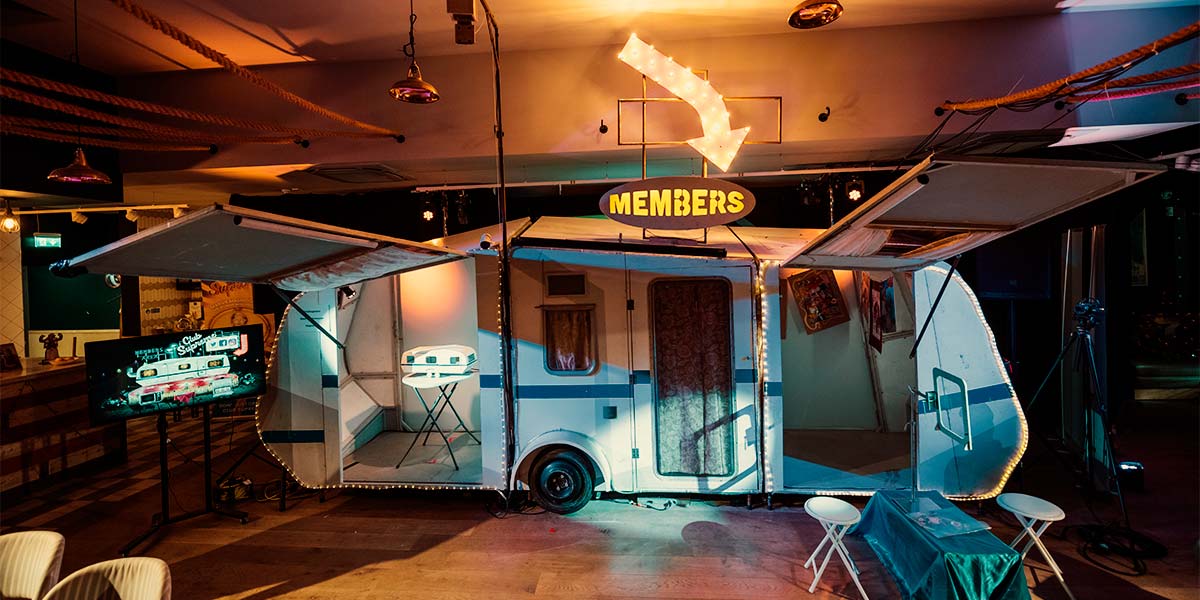 A white caravan sits inside a brightly lit room. Two portions of its side wall are opened upwards, revealing a tiny model of the same caravan and pictures hanging from a wall. A lit sign above the caravan says ‘Members’, and an arrow of lightbulbs points down at it. A large tv screen to the left shows a cartoon image of the caravan.
