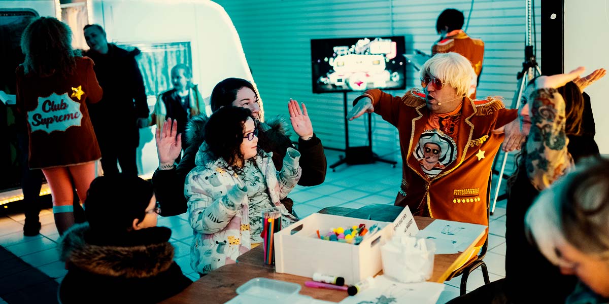 A group of children and adults sit around a table strewn with pens and paper. At the head sits a man wearing an orange jacket and sunglasses. The group hold their hands in the air at right angles to their bodies. Behind them, people mill around a caravan and television screen.
