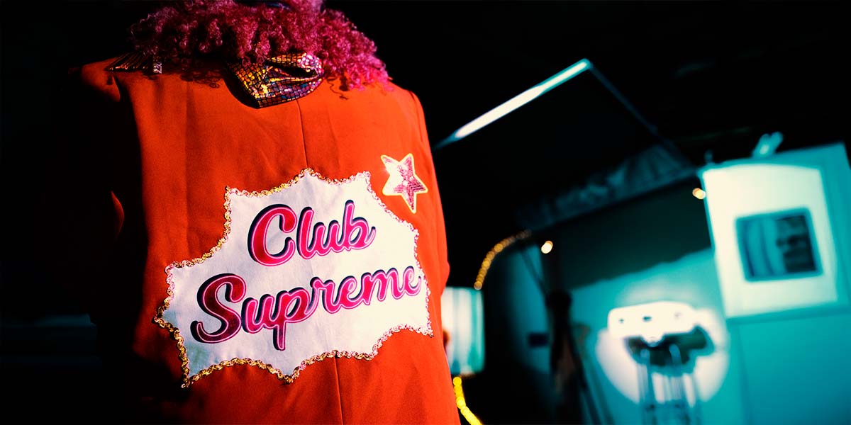 In red light, the back of an orange jacket with curly dark hair spilling over. at the top and the words Club Supreme printed in pink curly lettering over a white background. To the right and out of focus, a blue light glows over a tiny model of a caravan.
