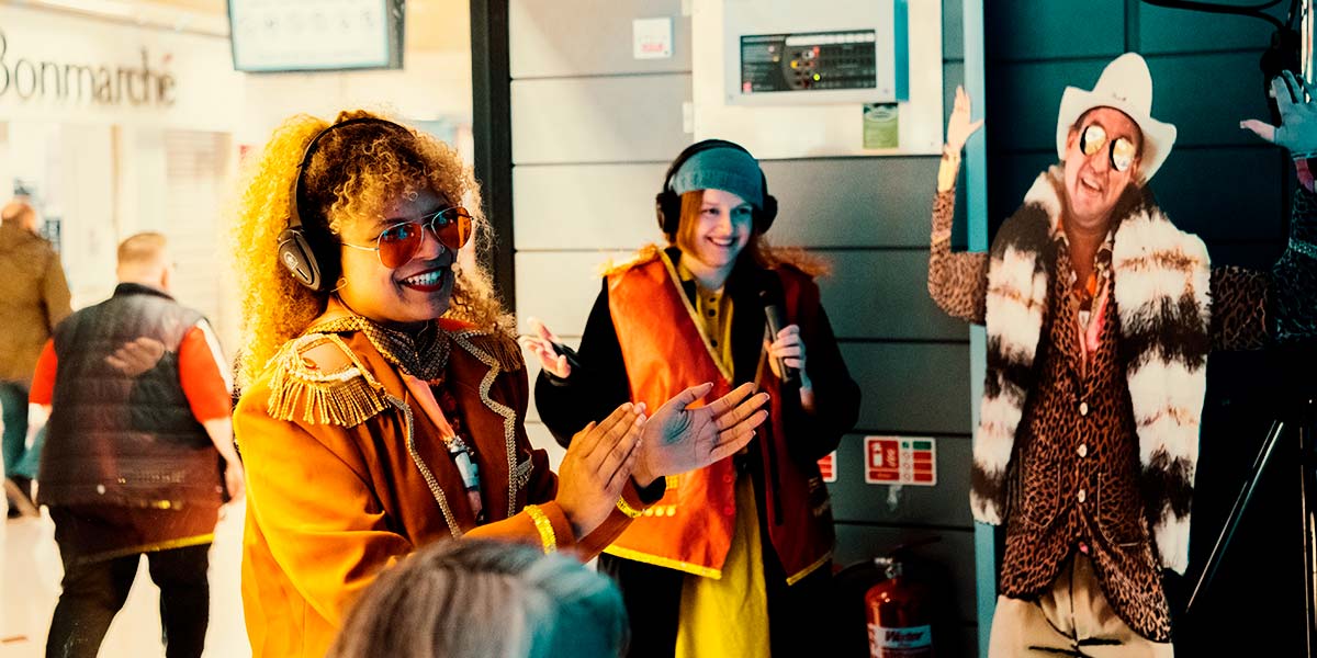  Inside a shop window, a grinning woman dressed in orange and wearing headphones claps her hands. Behind her, someone else in headphones smiles over a microphone they hold. Besides them is a cardboard cutout of a man in leopard print and furs, with his palms raised upwards.