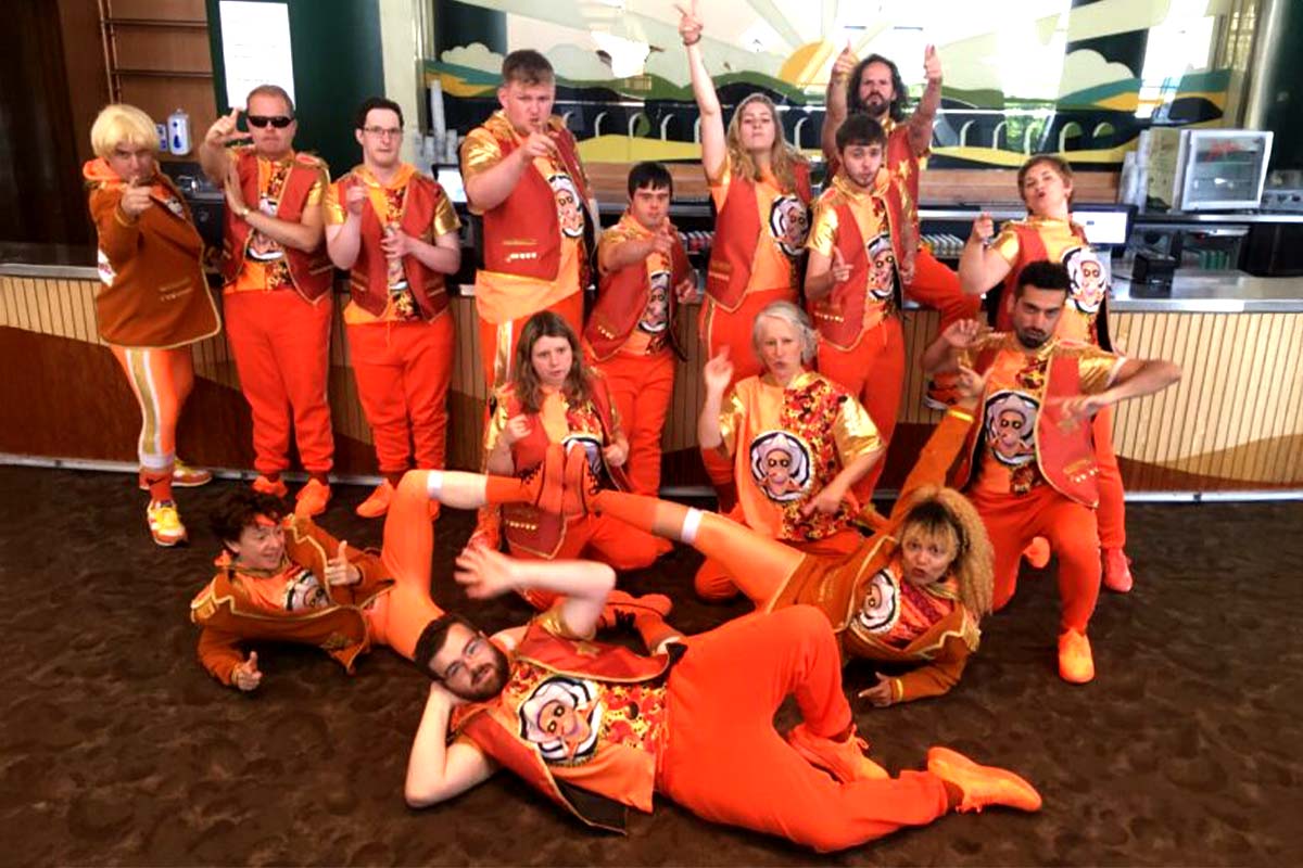 Fifteen people are dressed identically in bright orange trousers and shoes with open waistcoats over orange t-shirts bearing the print of a man wearing a Stetson and sunglasses. They pose in front of a stage, some lying on the floor, others standing and pointing to the camera.