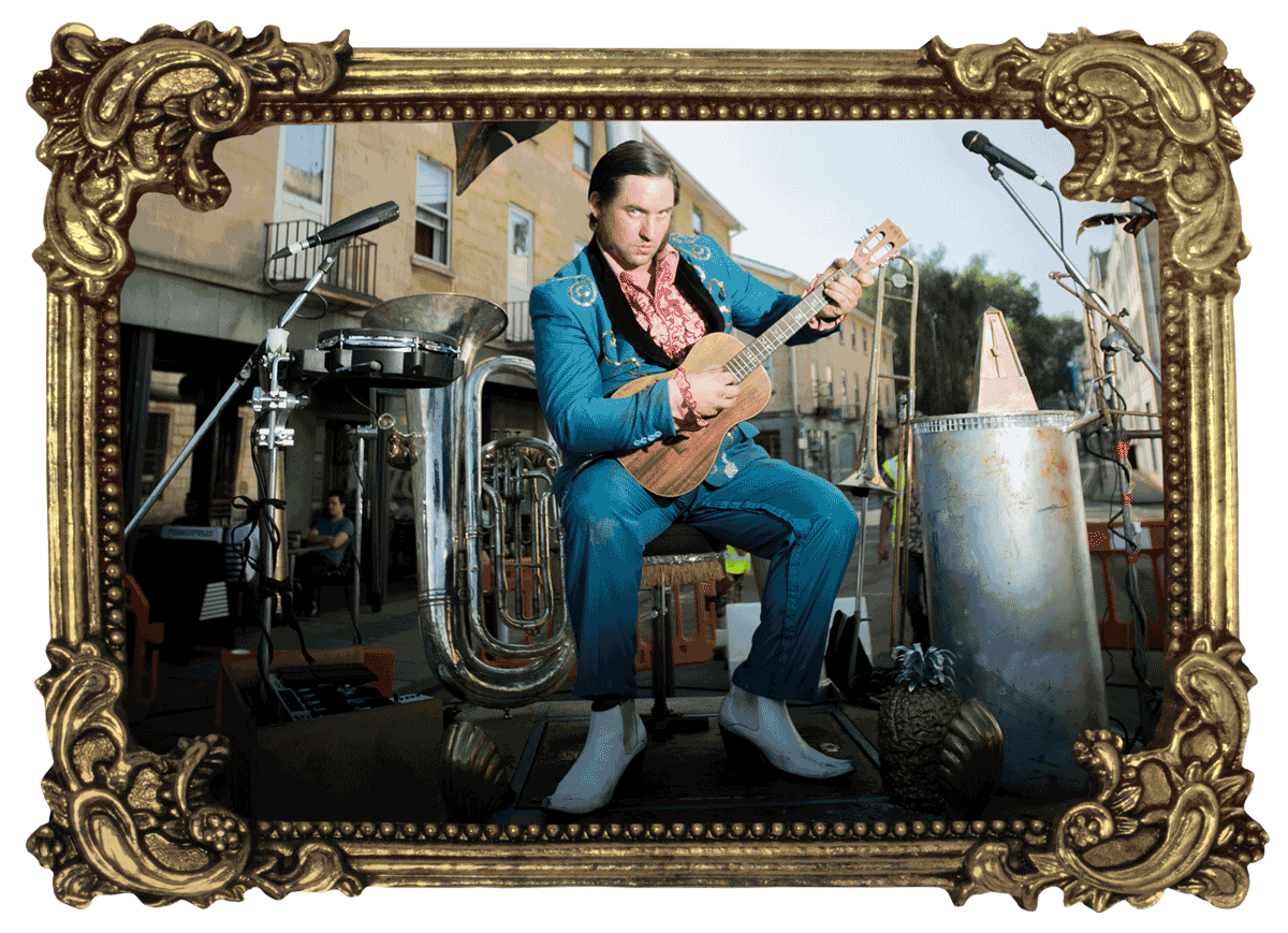 A man with slicked hair and suave electric blue suit poses with a baritone ukulele on a street. Clustered around him is an assortment of instruments and microphones whilst above, a metal palm tree opens its fronds to the sky.