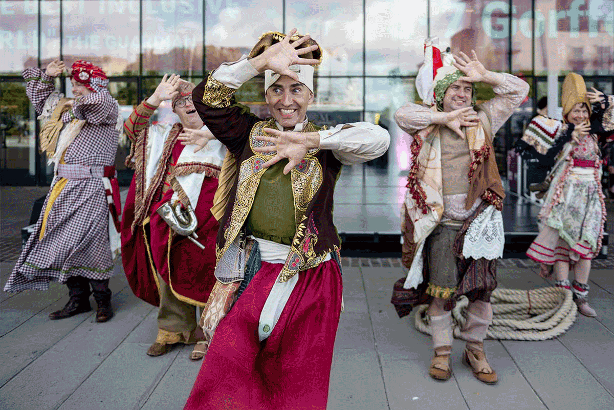 Five performers in colourful, tasselled costumes grin gleefully from behind their splayed fingers.

